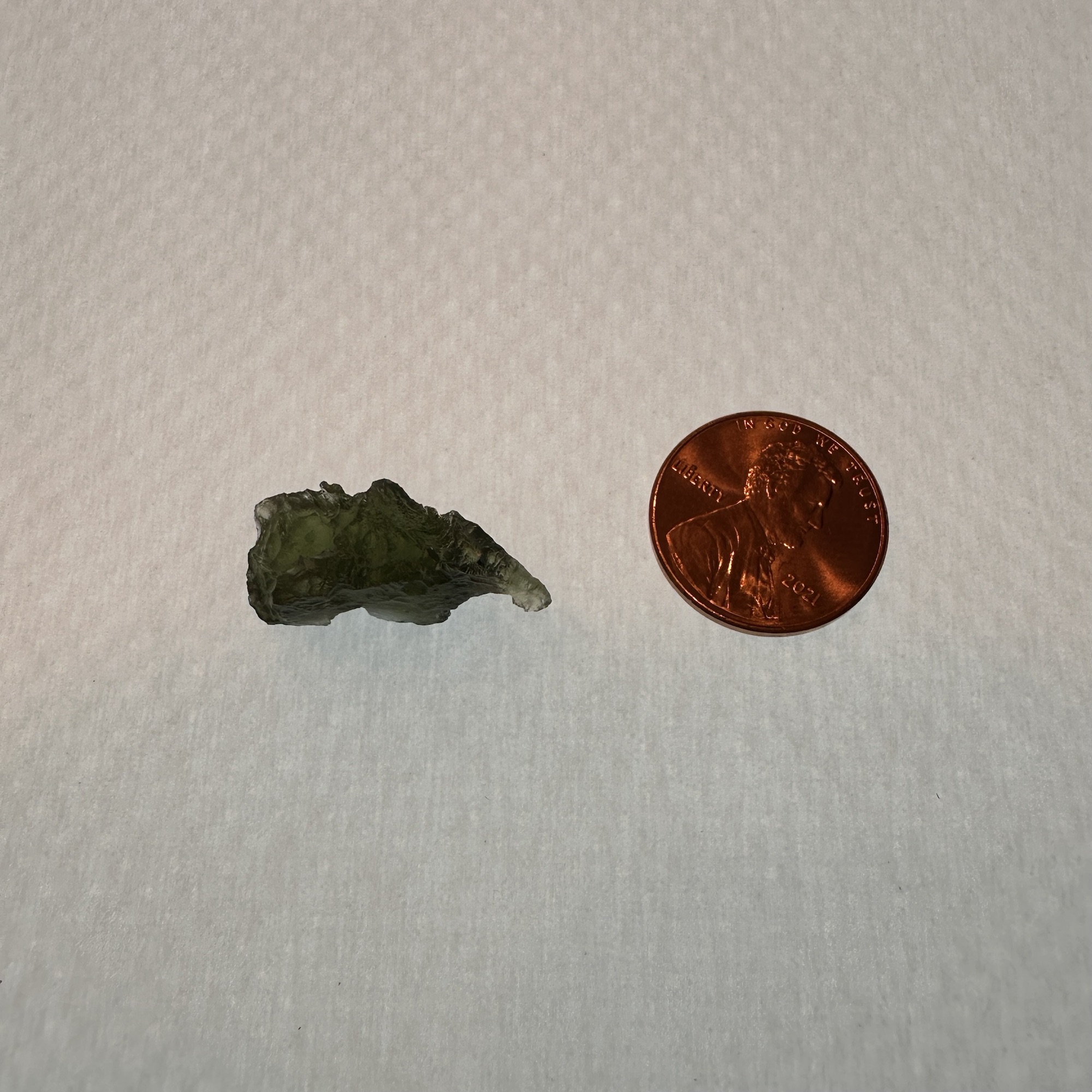 A green translucent Moldavite sitting next to a penny for a size comparison